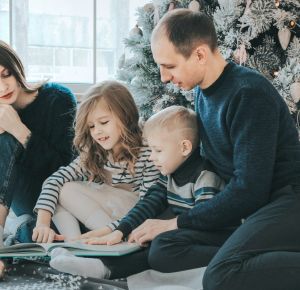 girl and boy reading book sitting between man and woman beside Christmas tree
